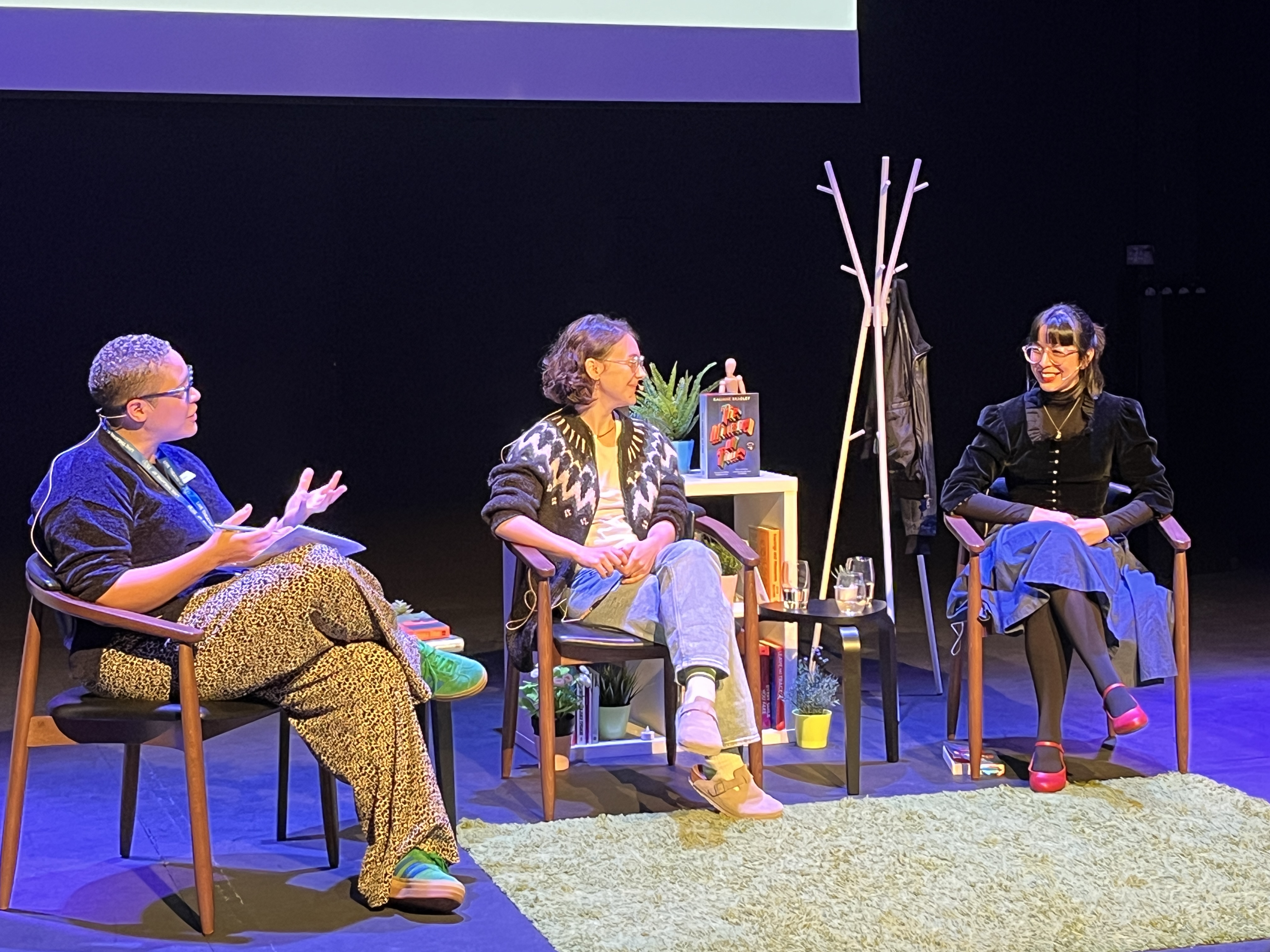 a black woman and two white presenting women sit on a stage, discuss and laugh